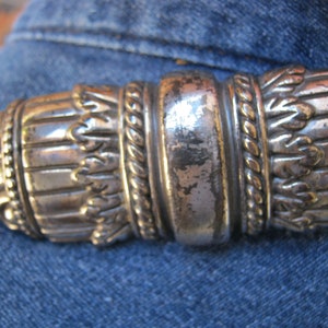 May include: A silver-toned metal belt buckle with intricate designs, including a rope-like pattern and a series of small, leaf-like shapes.