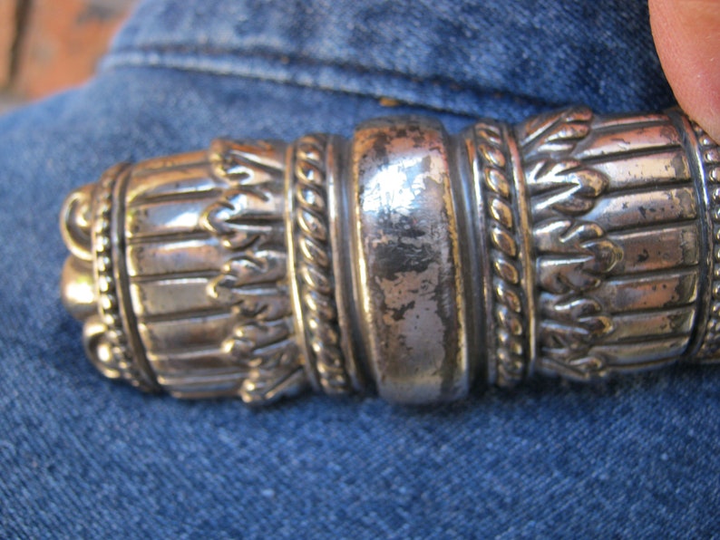 May include: A silver-toned metal belt buckle with intricate designs, including a rope-like pattern and a series of small, leaf-like shapes.