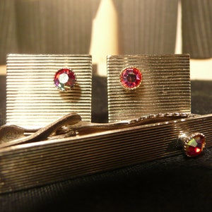 Op de afbeelding: Een set zilverkleurige manchetknopen en een stropdasklem. De rechthoekige manchetknopen en stropdasklem hebben een geribbeld ontwerp en zijn versierd met een centraal, iriserend, rond juweel. De set wordt gepresenteerd op een donkere ondergrond.