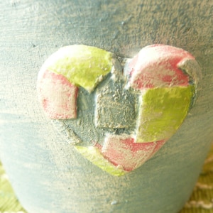 May include: A blue ceramic planter with a heart-shaped design. The heart is made of pink, green, and gray patchwork.