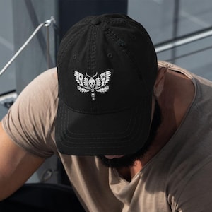 May include: A black baseball cap with a white embroidered moth design. The moth has a skull and crossbones on its body.
