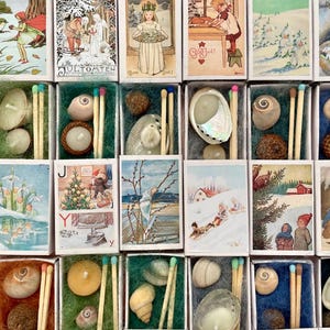Pode incluir: Uma coleção de caixas de fósforos, cada uma contendo uma pequena vela, conchas e fósforos. As caixas apresentam ilustrações vintage de cenas e personagens de inverno. As caixas estão dispostas em uma grade, mostrando a variedade de designs.
