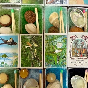 Pode incluir: Uma coleção de pequenas caixas de fósforos decoradas. Cada caixa contém um objeto diferente, como uma concha, uma bolota ou uma pequena vela. Algumas caixas apresentam ilustrações de cenas da natureza e contos de fadas. O texto "The Story of the Root Children" é visível.