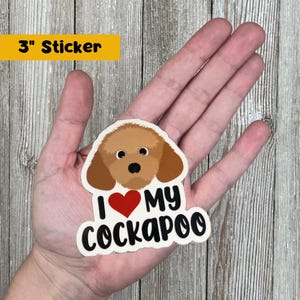 May include: A 3-inch sticker featuring a cartoon Cockapoo dog with brown fur, a red heart, and the text "I ❤️ MY COCKAPOO" in black and white. The sticker is held in a hand against a wooden background.