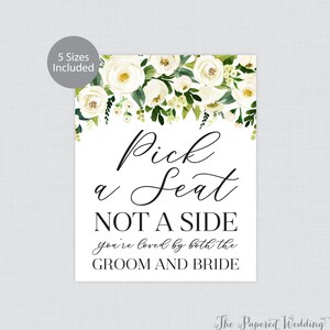 May include: A white wedding sign with floral accents and the text "Pick a Seat, NOT A SIDE, you're loved by both the GROOM AND BRIDE." The sign includes a floral border of white roses and green leaves.