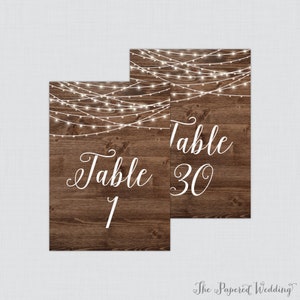 May include: Two rustic wooden table number signs with white string lights and the text "Table 1" and "Table 30".