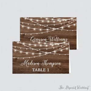 May include: Rustic wedding table card with a woodgrain background and white string lights. The card says "Grayson Williams" and "Madison Thompson" with "Table 1" below the names.