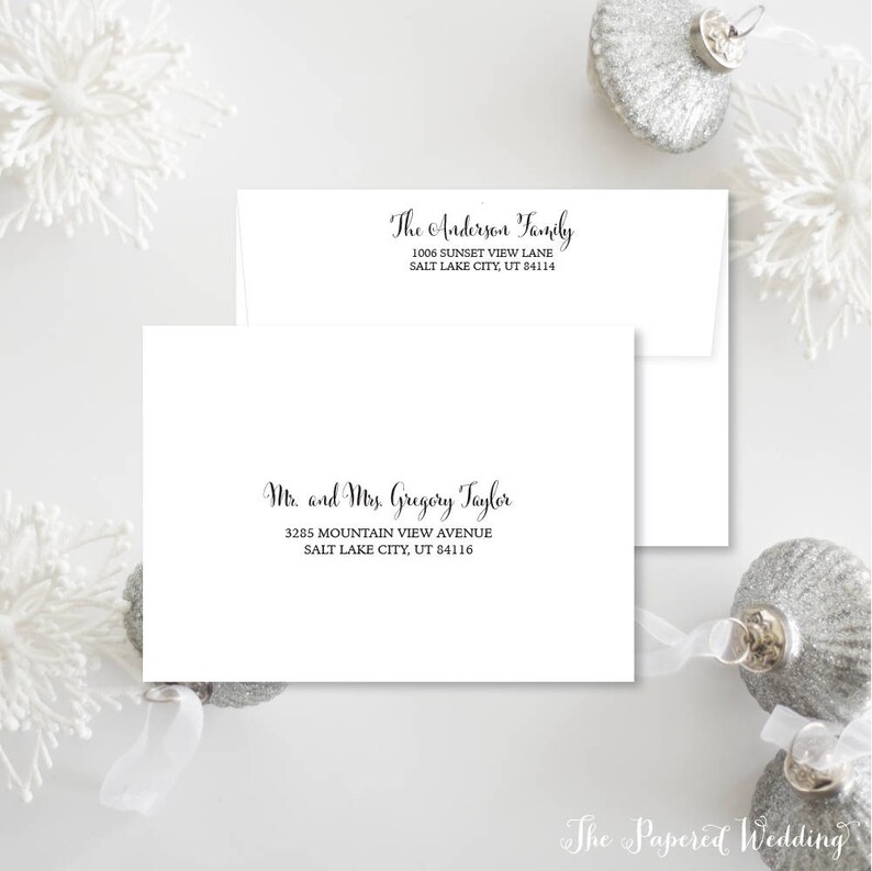 May include: White envelopes with black calligraphy. One envelope reads "The Anderson Family" with an address. The other reads "Mr. and Mrs. Gregory Taylor" with an address. The Papered Wedding logo is at the bottom. Silver ornaments and white floral accents are in the background.