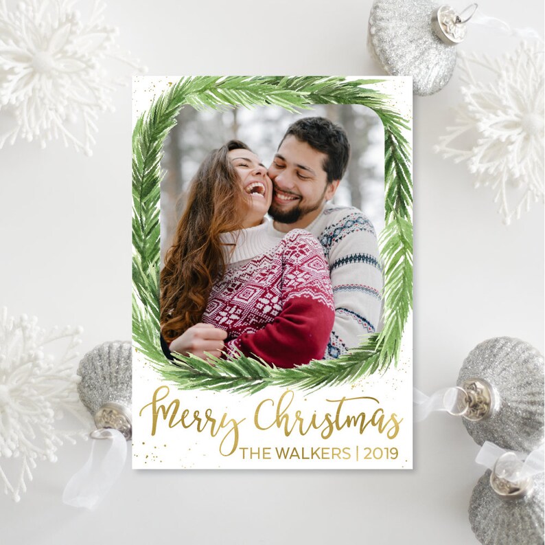 May include: A Christmas card featuring a couple laughing, framed by a green watercolor wreath. The words "Merry Christmas" are in gold script, with "The Walkers | 2019" below. Silver ornaments and snowflakes decorate the background.