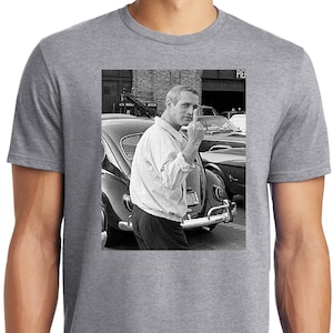 Puede incluir: Una foto en blanco y negro de un hombre haciendo el dedo medio mientras está de pie junto a un coche antiguo. El hombre lleva una camisa blanca abotonada y pantalones oscuros. El coche es un Volkswagen Beetle clásico.