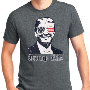 May include: A dark heather grey t-shirt with a black and white image of a man wearing sunglasses with an American flag design. The text "Trump Life" is printed in black below the image.