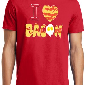 Puede incluir: Camiseta roja con el texto "Me encanta el bacon" en letras amarillas y naranjas. El coraz&oacute;n est&aacute; hecho de rayas de bacon y tiene un huevo frito encima.
