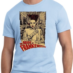 Puede incluir: Una camiseta azul claro con un dise&ntilde;o de p&oacute;ster vintage con la Novia de Frankenstein. El p&oacute;ster tiene el texto "The Bride of Frankenstein" y "Made from the skeletons of two women and the heart of a living girl."