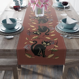 May include: A rectangular table runner in a deep terracotta color featuring a repeating pattern of black cats with yellow eyes and mouths open, interspersed with scattered autumn leaves in shades of gold, brown, and green, and small square geometric shapes.