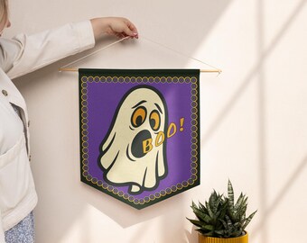 Può includere: Un arazzo a tema Halloween con un fantasma dei cartoni animati che dice "BOO!" in giallo. Il fantasma è bianco con grandi occhi arancioni e la bocca aperta. Lo stendardo è viola con un bordo verde e un'asta di legno nella parte superiore.