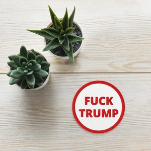 May include: Two small potted succulents and a round white patch with a red border. The patch has the words "FUCK TRUMP" in red, capital letters. The plants are green and in white pots.