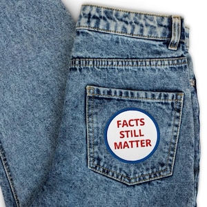 May include: Pale blue denim jeans with a patch on the back pocket. The patch is white with a blue border and the words "FACTS STILL MATTER" in red. The jeans have a classic five-pocket design.