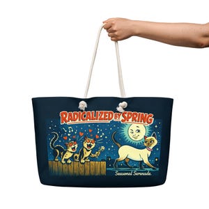 以下が含まれることがあります： 白いロープのハンドルが付いた、濃いネイビーのトートバッグ。バッグには、笑顔の月の下で、猫、ハート、音符の気まぐれなイラストが描かれています。バッグのテキストは「Radicalized by Spring」と「Seasonal Serenade」です。