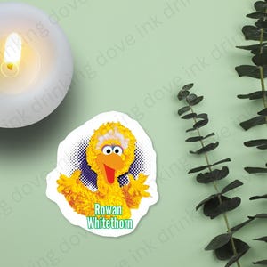 Peut inclure: Un autocollant jaune représentant Big Bird avec un bandeau blanc et le texte "Rowan Whitethorn".