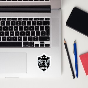 May include: A black and white sticker with a shield design. The shield has a dragon and a lion inside. The text "QUEST SQUAD" is on the shield.