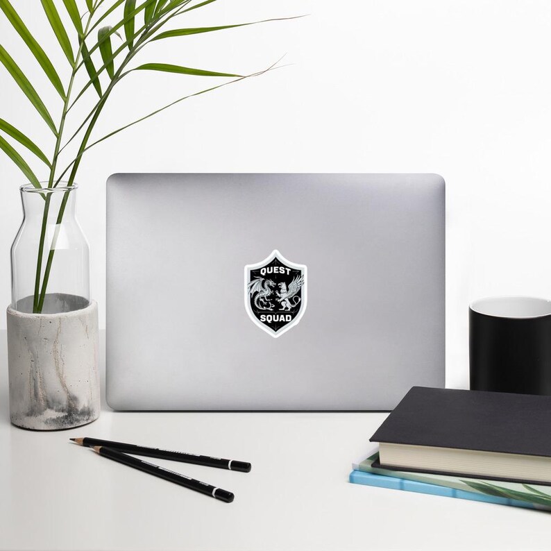 May include: A black and white sticker with a shield design. The shield has a dragon and a lion inside. The text "QUEST SQUAD" is written on the shield.