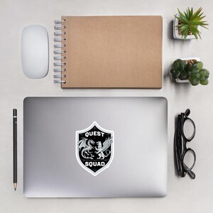 May include: Black and white sticker with a shield design. The shield has a dragon and a griffin facing each other. The text "QUEST SQUAD" is on the shield.