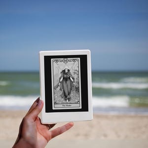 May include: A black and white tarot card with a woman in a long robe and a crown on her head. The card is labeled "The Healer" and has the letter "M" in the top right corner. The card is being held in front of a beach scene.