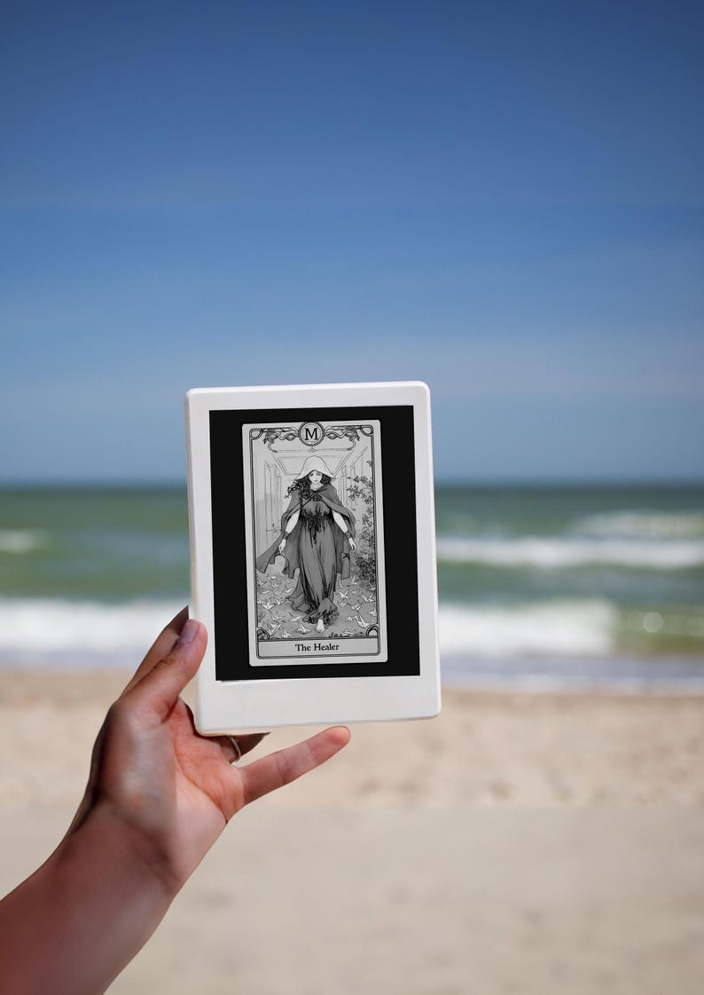 May include: A black and white tarot card with a woman in a long robe and a crown on her head. The card is labeled "The Healer" and has the letter "M" in the top right corner. The card is being held in front of a beach scene.