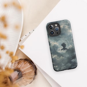 Könnte beinhalten: Smartphone-Hülle mit einer Drachensilhouette vor einem stürmischen Himmel. Die Hülle hat ein dunkelblaues und graues Farbschema mit einem kreisförmigen Designelement um den Drachen. Die Handyhülle liegt auf einer weißen Oberfläche.