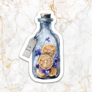 Op de afbeelding: Een blauwe glazen fles met een kurkstop, gevuld met gedroogde sinaasappelschijfjes en paarse bloemen. De fles heeft een etiket met de tekst "Jacky Denise".