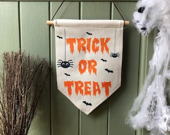 Pode incluir: Uma faixa de lona branca com o texto "Trick or Treat" em letras laranja. A faixa é decorada com aranhas e morcegos pretos.