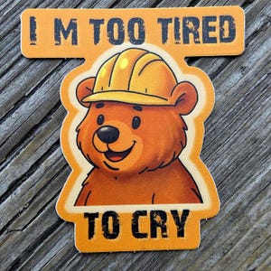 Op de afbeelding: Een oranje sticker met de tekst "I'M TOO TIRED TO CRY". De sticker toont een cartoonbeer met een gele helm. De beer is bruin met een zwarte neus en een lachende uitdrukking.