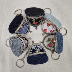 May include: Six small coin purses with metal kiss-lock frames and attached key rings. Each purse features a denim exterior in various shades of blue and black, with contrasting floral or patterned fabric linings in red, white, and green.