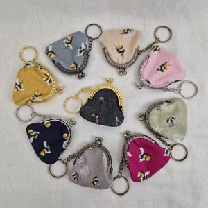 May include: Nine small coin purses with keychains, each featuring a different bee-themed fabric pattern. The purses are arranged in a circle on a light beige surface.