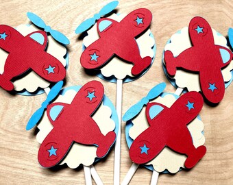 Puede incluir: Cinco aviones de papel rojos con hélices azules y estrellas blancas sobre un fondo beige. Los aviones están unidos a palos blancos.