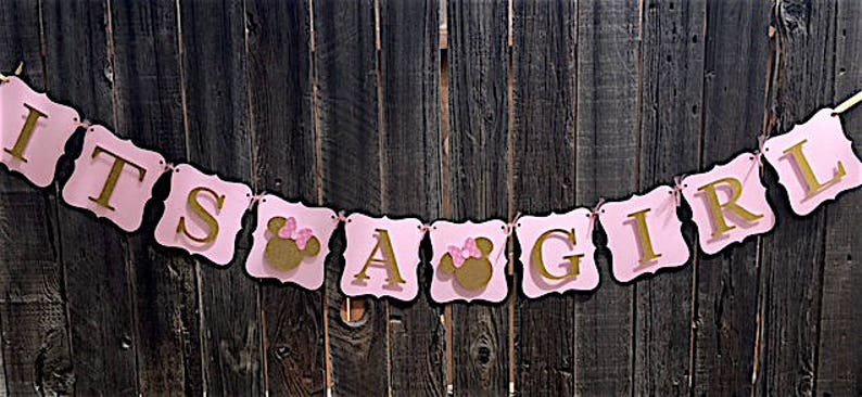Puede incluir: Bandera rosa y dorada con el texto "It's a Girl" y una cabeza de Minnie Mouse en cada una de las letras "A" y "G".