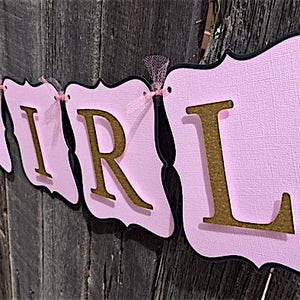 Puede incluir: Bandera rosa y negra con letras doradas de purpurina que deletrean "Baby Girl".
