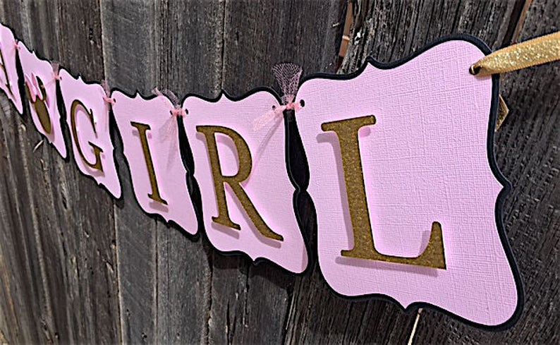 Puede incluir: Bandera rosa y negra con letras doradas de purpurina que deletrean "Baby Girl".