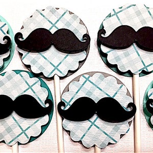 Op de afbeelding: Set van zes cupcake toppers met een zwarte snor op een blauw-wit geruite achtergrond. De toppers zijn bevestigd aan witte stokjes.