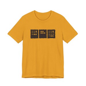 May include: Mustard yellow t-shirt featuring a dark brown graphic of three rectangular shapes. Each shape has a different arrangement of small, horizontal lines and squares, creating a minimalist design. The shirt is made of a soft material.