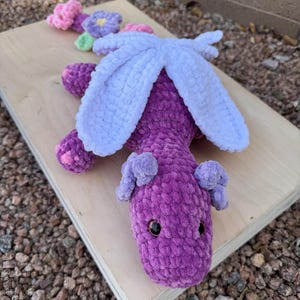 Puede incluir: Un drag&oacute;n de ganchillo morado hecho a mano con alas azul claro y peque&ntilde;as orejas moradas. El drag&oacute;n tiene ojos de seguridad negros y est&aacute; sobre una superficie de madera. Flores de ganchillo en rosa, morado y verde est&aacute;n junto al drag&oacute;n.