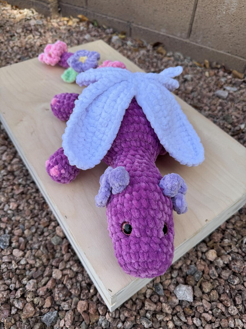 Puede incluir: Un drag&oacute;n de ganchillo morado hecho a mano con alas azul claro y peque&ntilde;as orejas moradas. El drag&oacute;n tiene ojos de seguridad negros y est&aacute; sobre una superficie de madera. Flores de ganchillo en rosa, morado y verde est&aacute;n junto al drag&oacute;n.