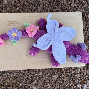 Puede incluir: Un drag&oacute;n morado de peluche hecho a mano con un ala azul claro y detalles florales. El drag&oacute;n est&aacute; decorado con flores rosas, amarillas y moradas. El drag&oacute;n est&aacute; sobre una superficie de madera.