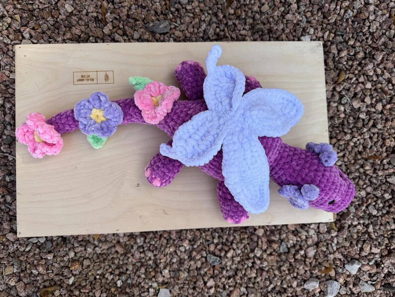 Puede incluir: Un drag&oacute;n morado de peluche hecho a mano con un ala azul claro y detalles florales. El drag&oacute;n est&aacute; decorado con flores rosas, amarillas y moradas. El drag&oacute;n est&aacute; sobre una superficie de madera.