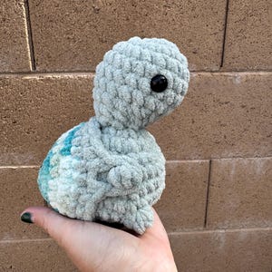 May include: A handmade, light blue crochet turtle with a black eye. The turtle has a textured, plush appearance and a small, rounded body. The turtle is held in a hand against a brick wall background.