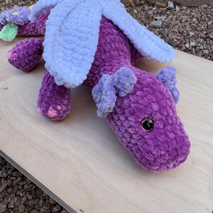Puede incluir: Un drag&oacute;n de ganchillo morado hecho a mano con alas blancas y peque&ntilde;as orejas moradas. El drag&oacute;n tiene una flor con detalles verdes y morados en la cola. El drag&oacute;n tiene ojos de seguridad negros y est&aacute; sobre una superficie de madera.