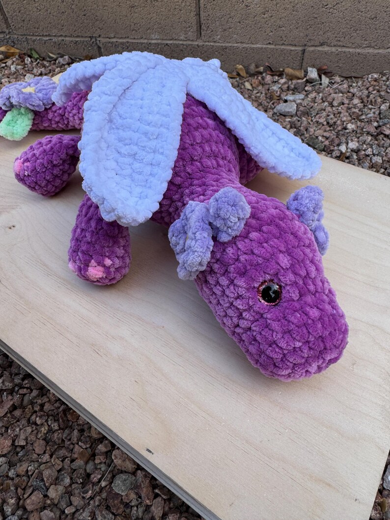 Puede incluir: Un drag&oacute;n de ganchillo morado hecho a mano con alas blancas y peque&ntilde;as orejas moradas. El drag&oacute;n tiene una flor con detalles verdes y morados en la cola. El drag&oacute;n tiene ojos de seguridad negros y est&aacute; sobre una superficie de madera.