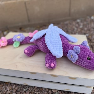 Puede incluir: Un drag&oacute;n de ganchillo morado hecho a mano con alas azul claro y detalles de flores rosas y moradas. El drag&oacute;n est&aacute; sobre una superficie de madera clara, con un fondo borroso de rocas y una pared.