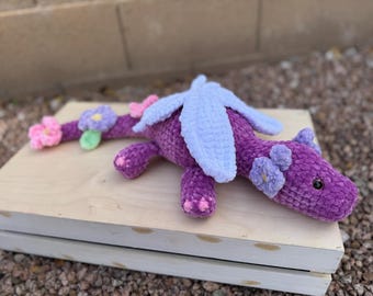May include: A handmade purple crochet dragon with light blue wings and pink and purple flower accents. The dragon is lying on a light-colored wooden surface, with a blurred background of rocks and a wall.