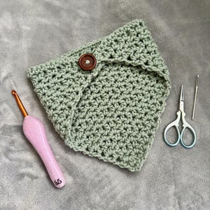 Peut inclure: Une pochette crochetée vert sauge avec une fermeture à bouton en bois. Un crochet rose, de petits ciseaux et une aiguille sont à côté de la pochette. Le crochet est de taille 5,5.